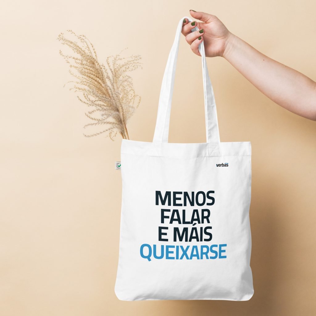 Una mano sostiene una bolsa de tela con la frase MENOS FALAR E MAIS QUEIXARSE en una pared de color beige.