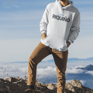 Un hombre sonriendo con ropa de invierno y gorro, está de pie en una montaña con un cielo claro y nubes abajo.